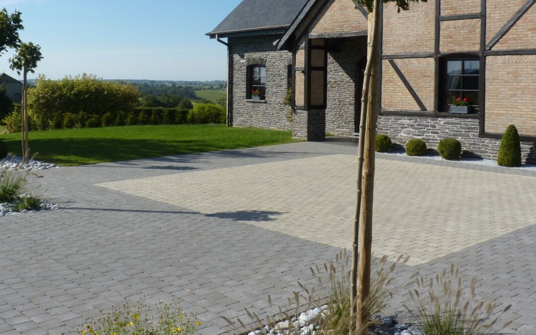 Une grande cour pavée en béton gris et beige, devant une maison en pierre et brique