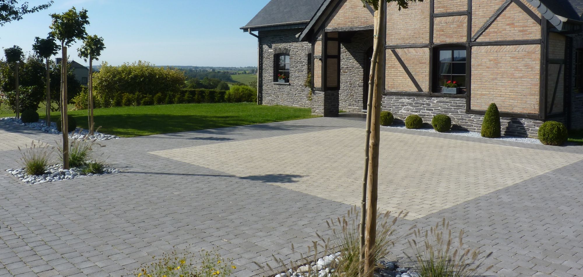 Une grande cour pavée en béton gris et beige, devant une maison en pierre et brique