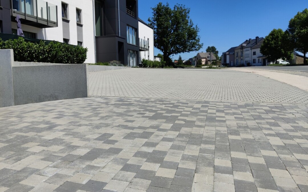 Vue d'une allée pavée moderne dans un quartier résidentiel. Les pavés carrés sont disposés en motifs de différentes teintes de gris, créant un contraste élégant avec l'environnement. La surface de l'allée est divisée en deux zones : une partie en pavés uniformes au premier plan et une partie en pavés avec des interstices pour le drainage à l'arrière-plan.